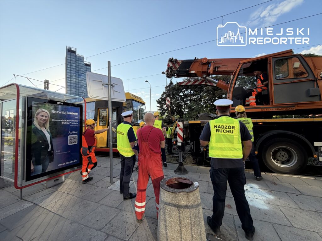Grupa pracowników w odzieży roboczej oraz policjantów obserwuje akcję naprawy wykolejonego tramwaju przy uszkodzonym torowisku.