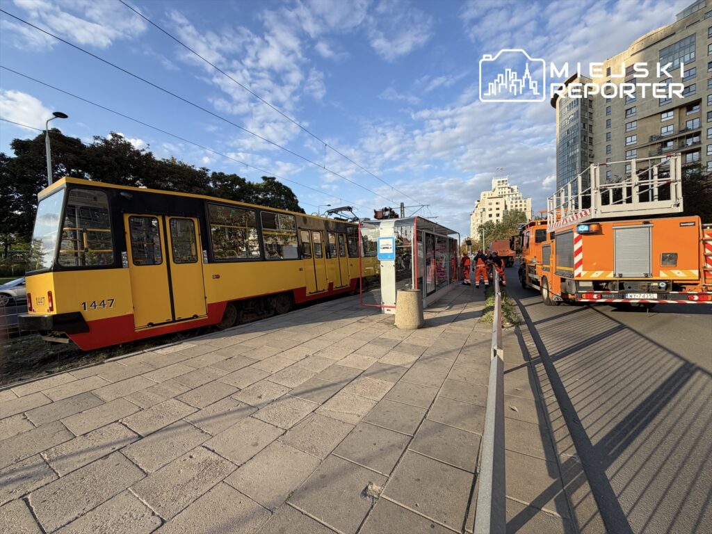 Wykolejony tramwaj w żółto-czerwonym kolorze stoi obok przystanku, a ratownicy w pomarańczowych uniformach pracują przy uszkodzonym torowisku.