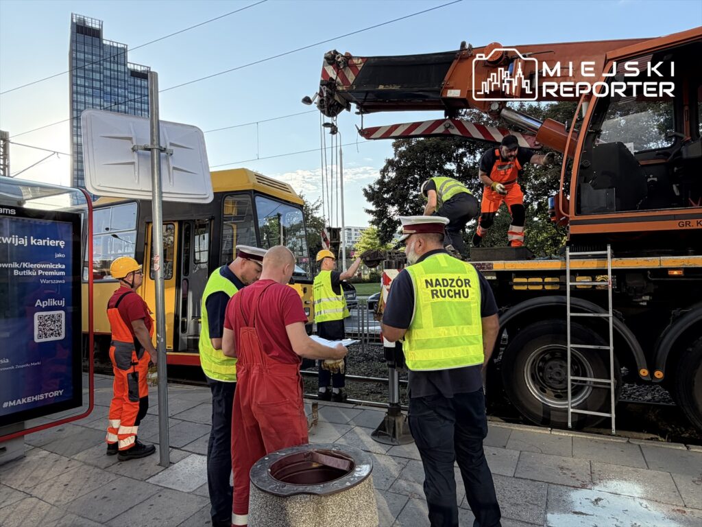 Grupa pracowników w odblaskowych kamizelkach naprawia uszkodzone torowisko tramwajowe, obok wykolejonego tramwaju.