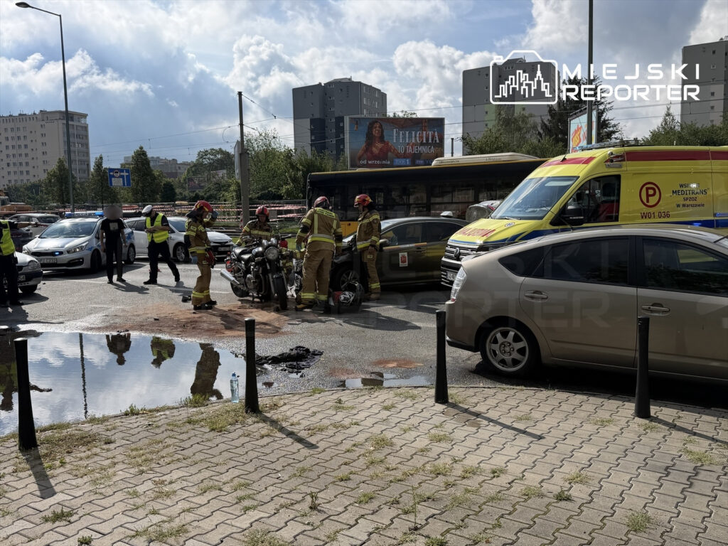Strażacy badają miejsce wypadku drogowego, gdzie leży motocykl, a obok znajduje się karetka i policyjne pojazdy.