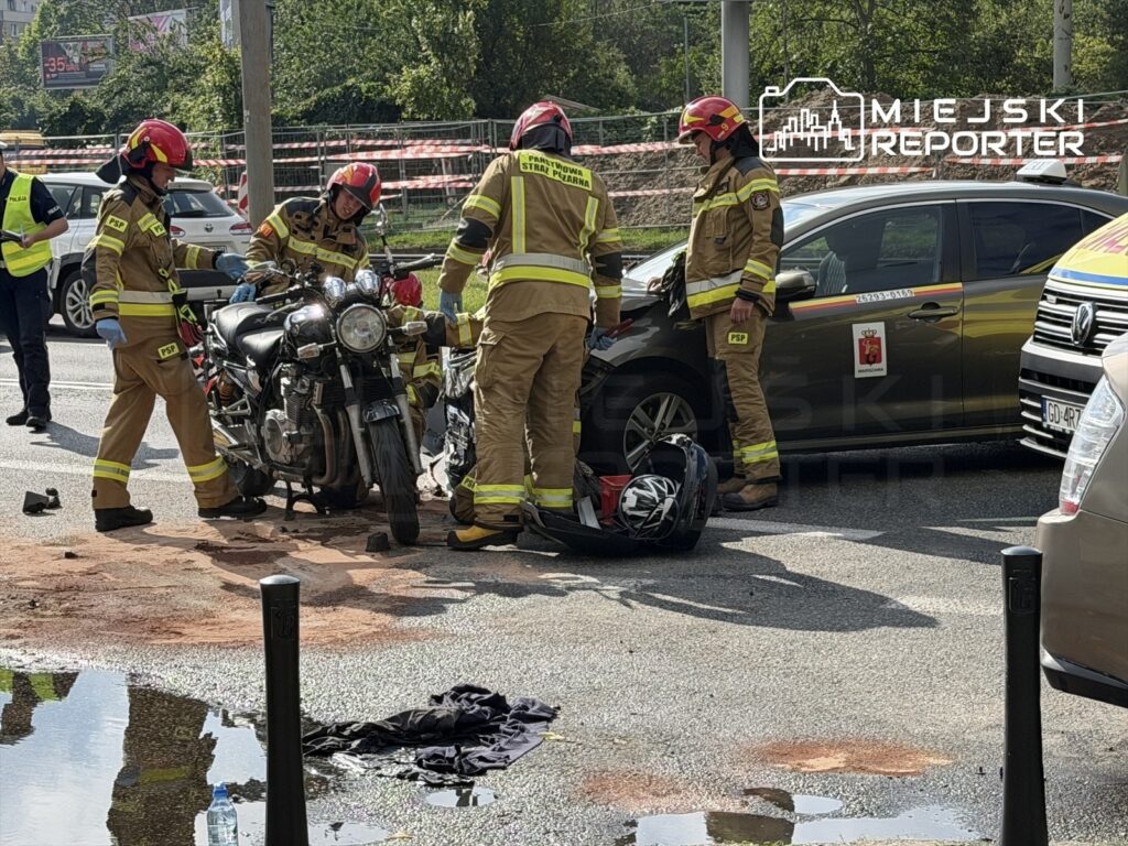Czterech strażaków w mundurach zabezpiecza miejsce wypadku motocyklowego na ulicy, obok leżącego motocykla.