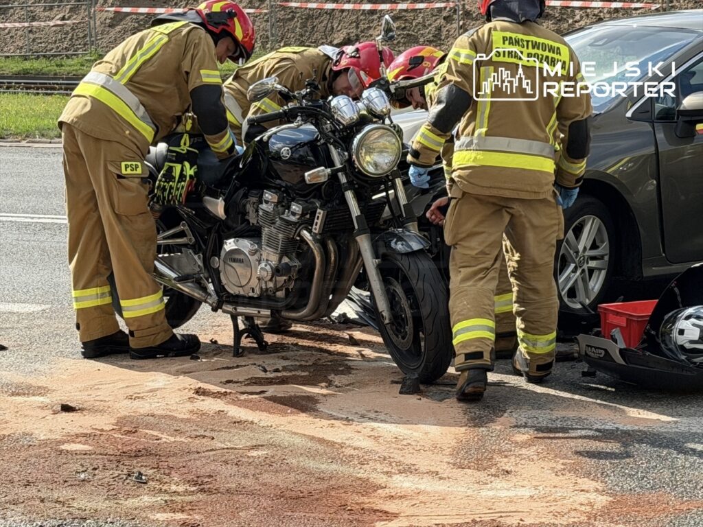 Strażacy w mundurach zabezpieczają motocykl na drodze po wypadku, obok widoczna plama oleju.