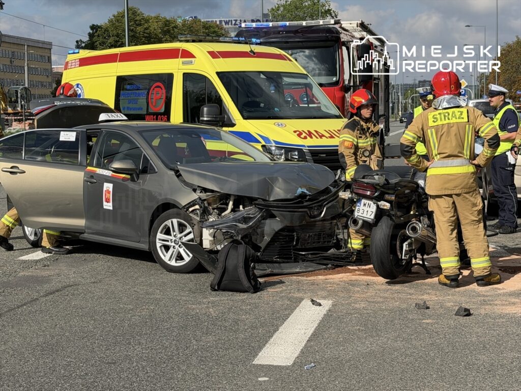 Wypadek drogowy z udziałem samochodu osobowego i motocykla, w tle strażacy oraz karetka pogotowia.