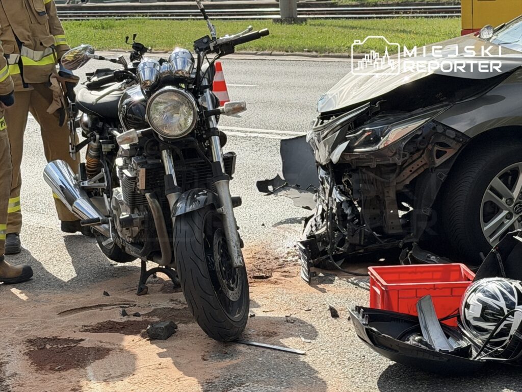 Motocykl leży na drodze obok uszkodzonego samochodu, a strażak w mundurze bada sytuację. W tle widać ruch uliczny.