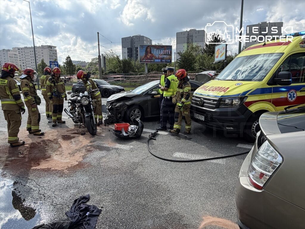 Grupa strażaków w mundurach ocenia skutki wypadku drogowego z udziałem samochodu osobowego i motocykla na ruchliwej ulicy.