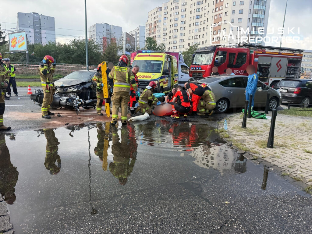 Grupa ratowników medycznych i strażaków udziela pomocy poszkodowanemu na miejscu wypadku drogowego.