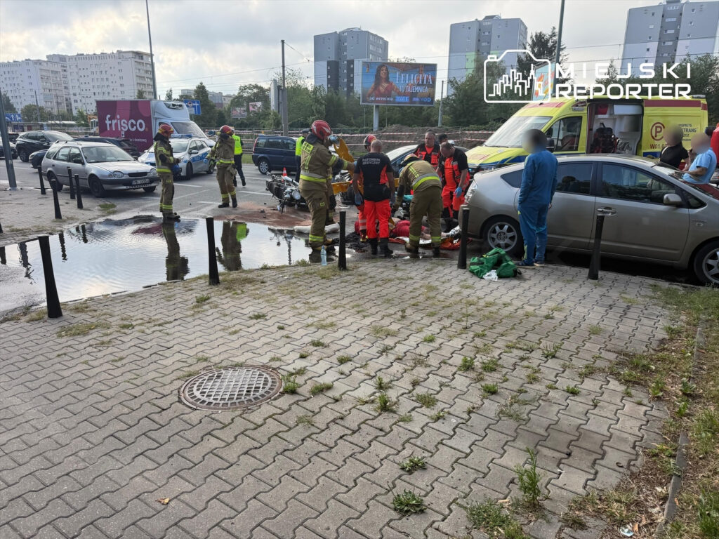 Grupa ratowników medycznych i strażaków udziela pomocy poszkodowanemu na chodniku obok zalanej wodą jezdni.