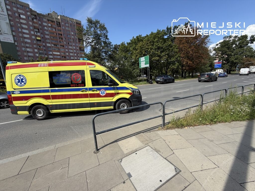Karetka pogotowia ratunkowego przejeżdżająca obok ruchliwej ulicy, w tle widać zieleń drzew i budynki mieszkalne.