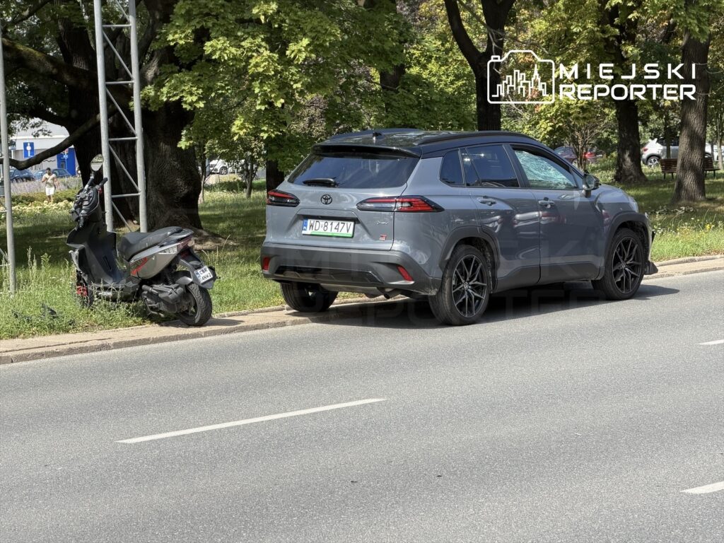 Szary SUV zaparkowany obok uszkodzonego motoroweru przy drodze, w tle zieleń drzew i ławki.