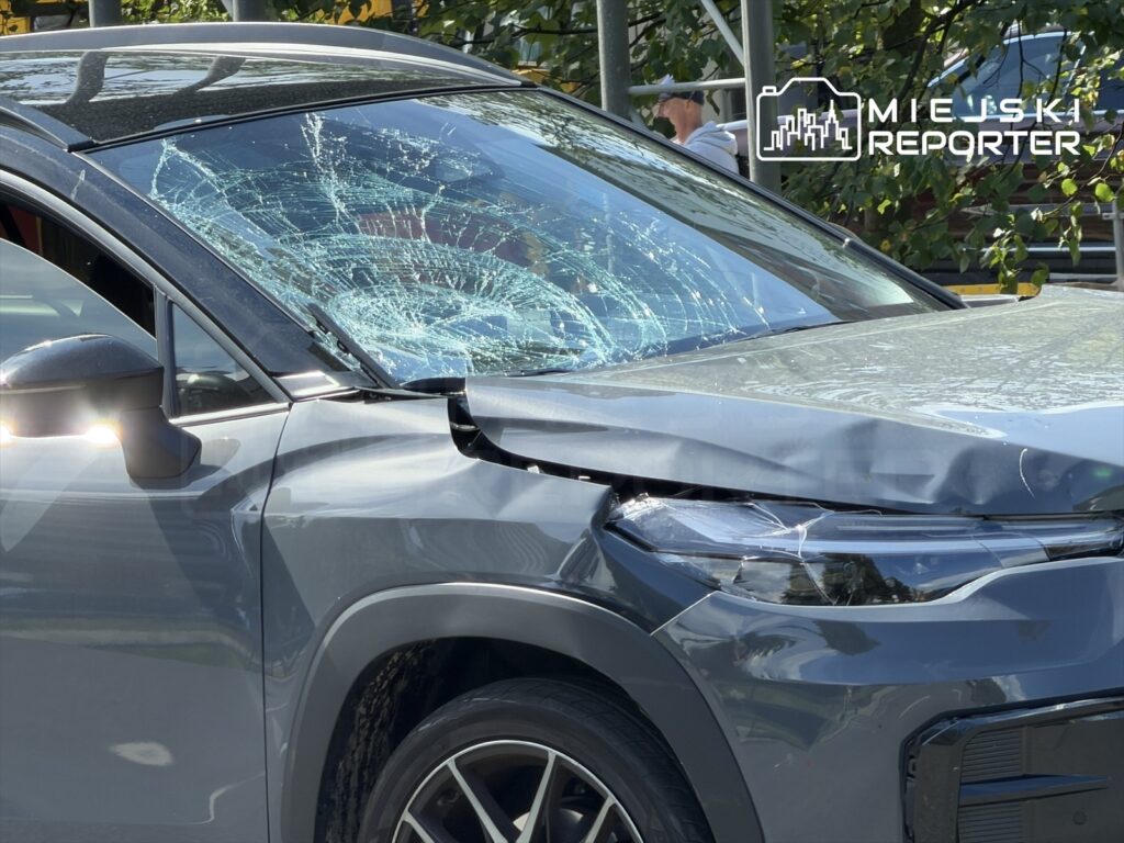 Uszkodzony samochód osobowy z wybitym przednim szybą, z widocznymi wgnieceniami na masce, zaparkowany w miejskim otoczeniu.