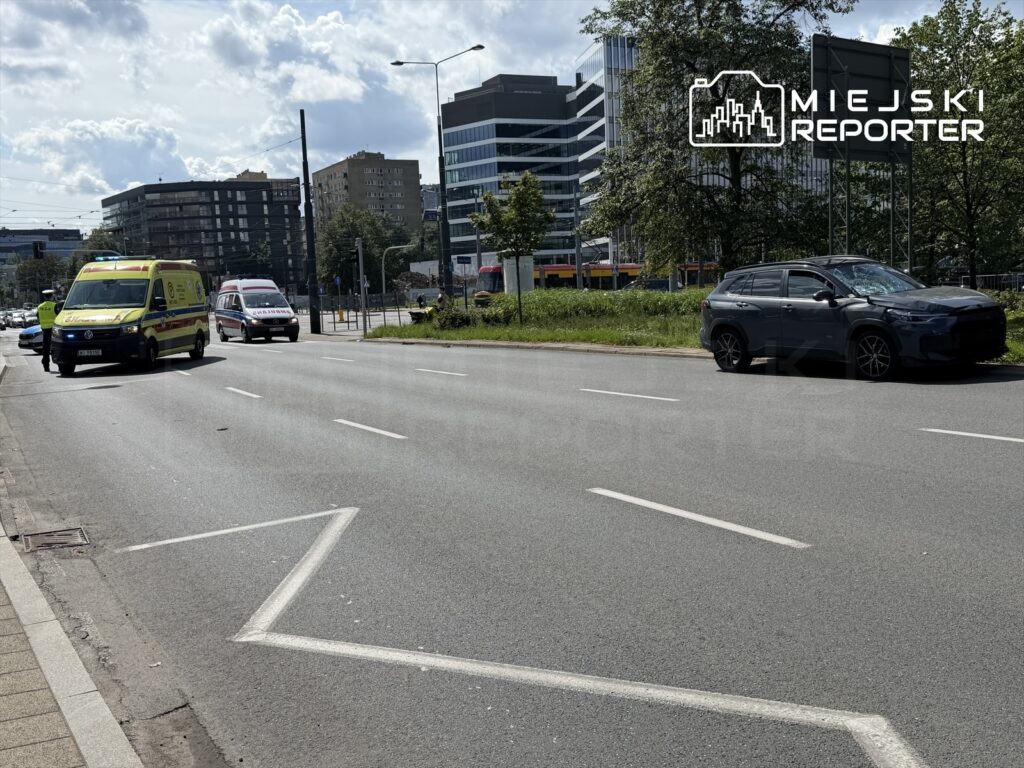 Karetkę pogotowia i radiowóz zatrzymane na jezdni obok uszkodzonego samochodu w miejskim otoczeniu.