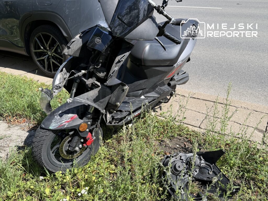 Zniszczony motorower leży na trawie obok asfaltowej drogi, z widocznymi uszkodzeniami przodu i odłamkami obok.