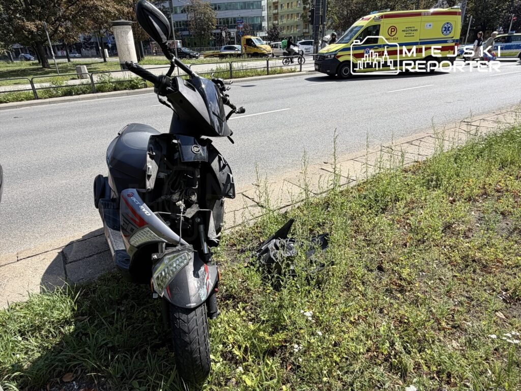 Uszkodzony motorower stoi przy krawężniku, obok ruchliwej ulicy, z widocznymi uszkodzeniami. W tle przejeżdża ambulans.
