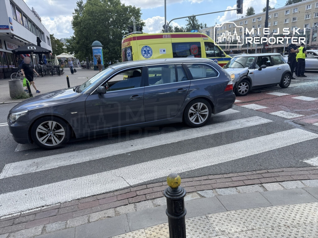 Samochód osobowy zderzył się z innym pojazdem na przejściu dla pieszych, w tle widać ambulans oraz przechodniów.