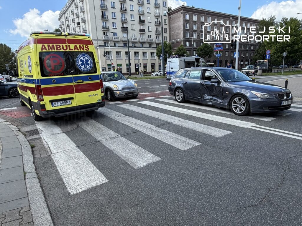 Karetka pogotowia zatrzymana na przejściu dla pieszych obok uszkodzonego samochodu osobowego w mieście.