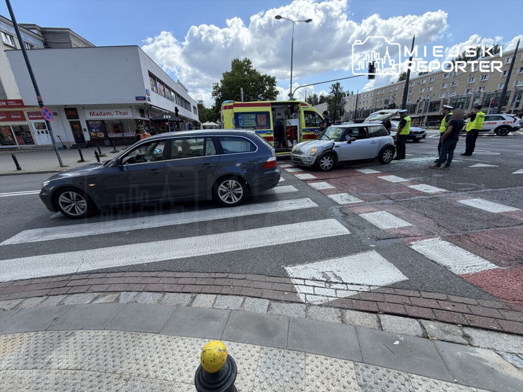Dwa samochody, w tym szary pojazd i mały samochód osobowy, zderzone na przejściu dla pieszych, obok przybyła karetka i policja.