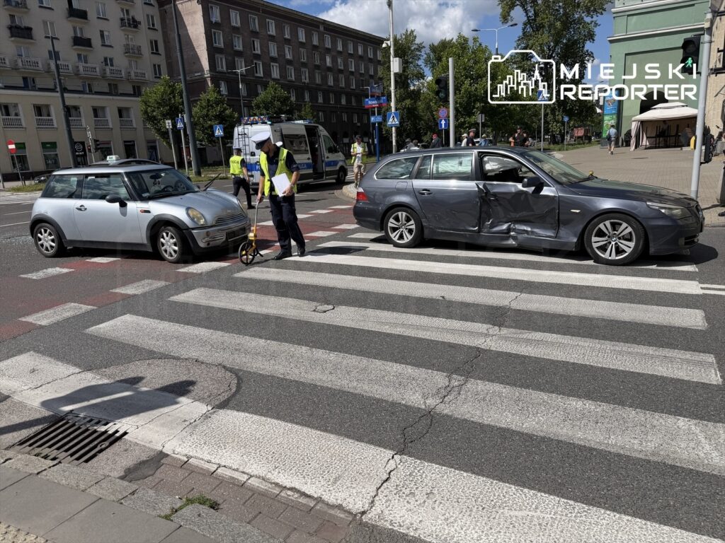 Policjant mierzy odległości na pasach, obok uszkodzony samochód osobowy oraz taksówka na skrzyżowaniu.