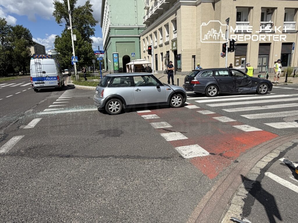 Dwa uszkodzone samochody na przejściu dla pieszych, obok policyjny radiowóz oraz grupa ludzi obserwujących zdarzenie.