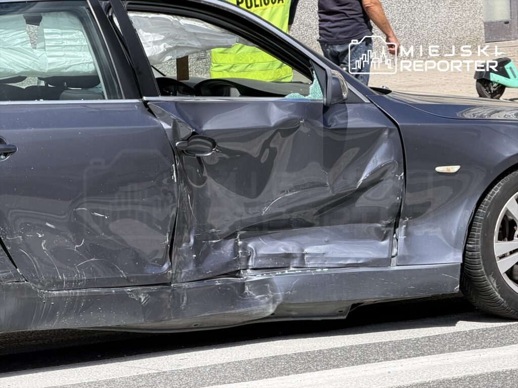 Uszkodzona lewa strona czarnego samochodu osobowego z wgniecioną drzwiami, stojącego na ulicy obok policjantów.