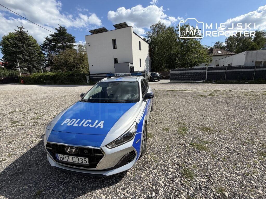 Policyjny samochód zaparkowany na utwardzonej nawierzchni w otoczeniu budynków mieszkalnych i zieleni.