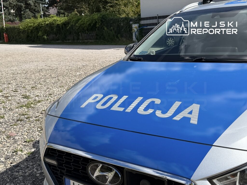 Nieoznakowany radiowóz policyjny zaparkowany na żwirowym placu, z widoczną nazwą "POLICJA" na dachu.