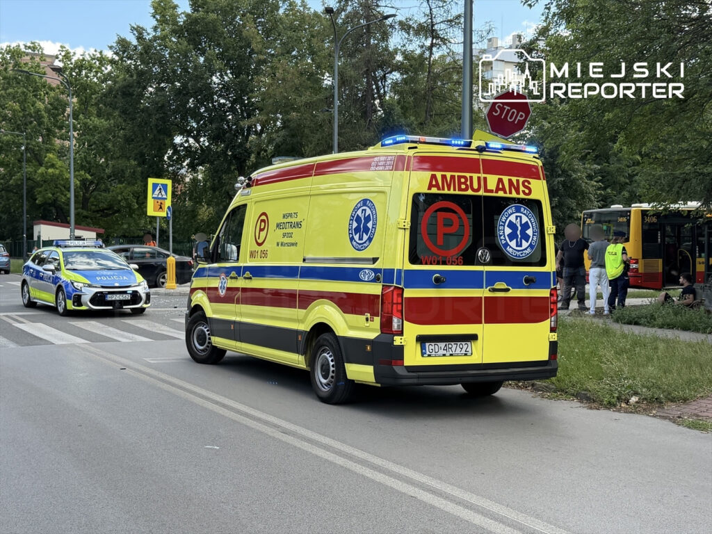 Karetka pogotowia i radiowóz policyjny na skrzyżowaniu, obok grupy osób i autobusu stojącego na poboczu.