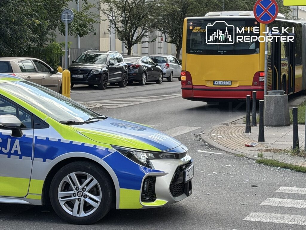Policyjny radiowóz zatrzymany przy ulicy, obok żółty autobus komunikacji miejskiej, w tle zaparkowane samochody.