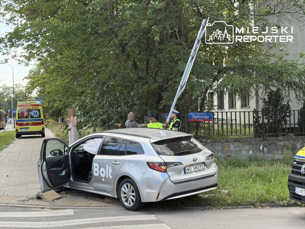 W pobliżu uszkodzonego samochodu marki Toyota z logo "Bolt" stoi kobieta, a obok interweniują służby ratunkowe.