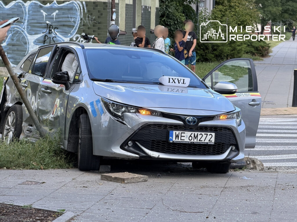 Rozbita srebrna Toyota leży przy krawężniku, z uszkodzoną stroną i otwartymi drzwiami, obok grupy dzieci.