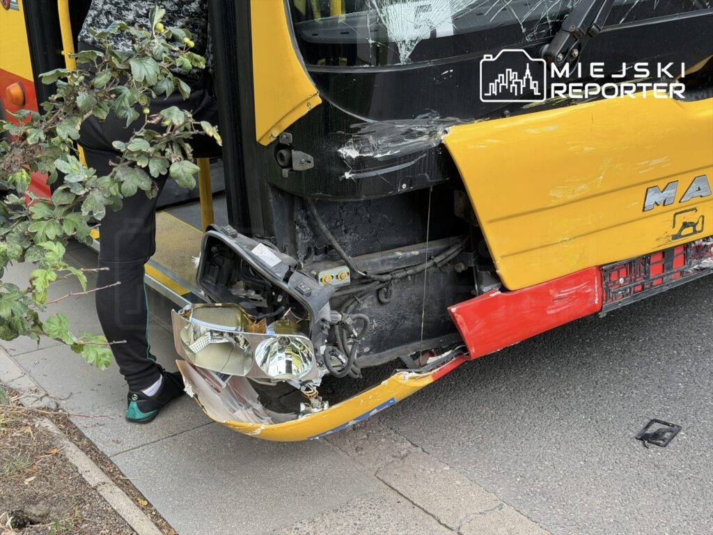 Zniszczony autobus z uszkodzoną przednią częścią stoi na ulicy, obok rosnące krzewy.