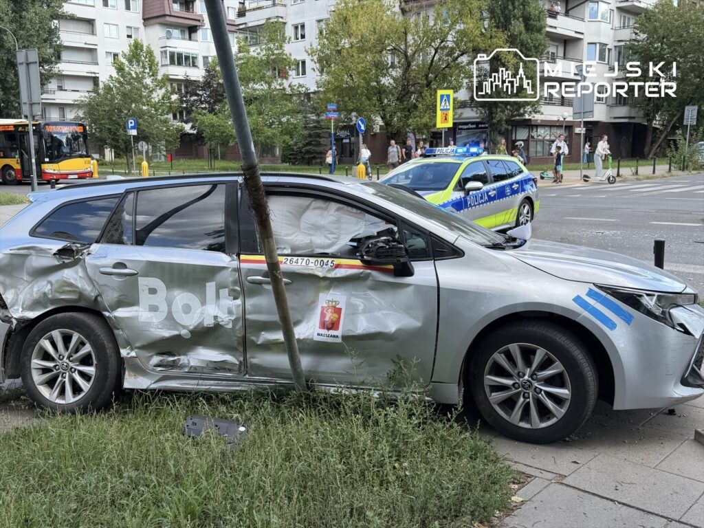 Uszkodzony samochód osobowy z logiem Bolt stoi na poboczu, obok policyjny radiowóz oraz przechodnie obserwujący zdarzenie.