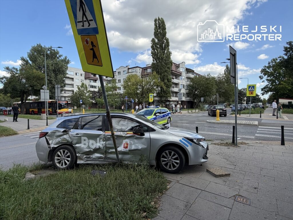 Zniszczony samochód osobowy z logo "Bolt" stoi na poboczu, obok uszkodzonego znaku drogowym, w tle widać autobus i ludzi na chodniku.
