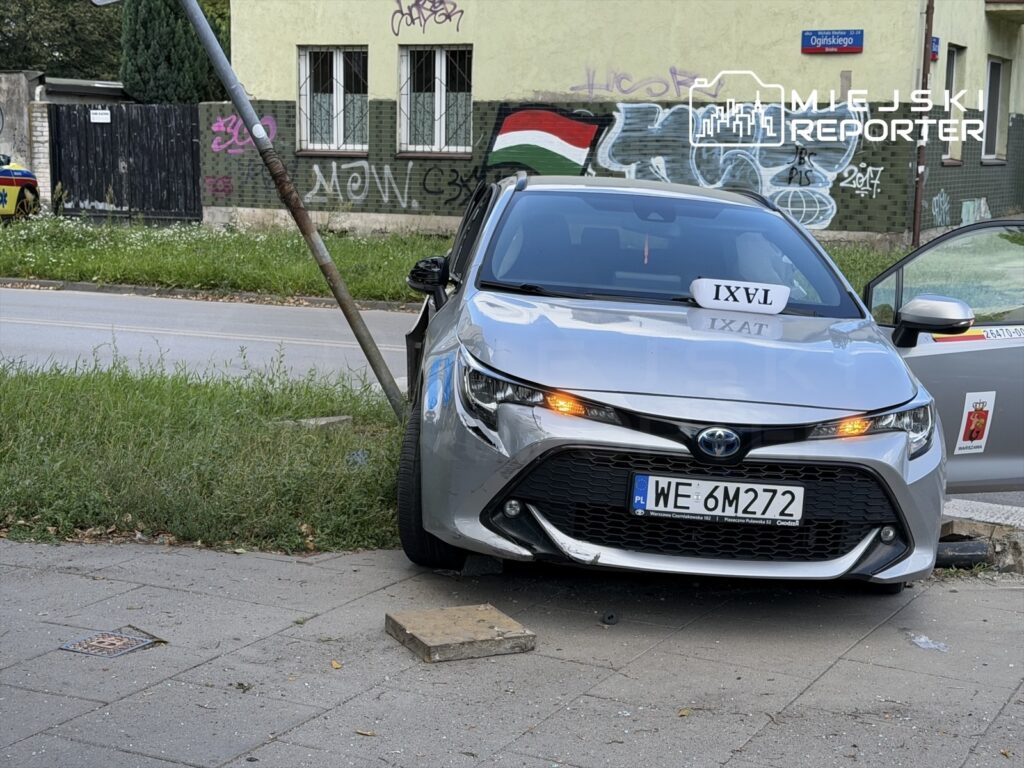 Toyota z oznaczeniem "TAXI" stoi na chodniku z uszkodzoną przednią częścią, obok zgiętego słupa. W tle widoczna graffiti na ścianie budynku.
