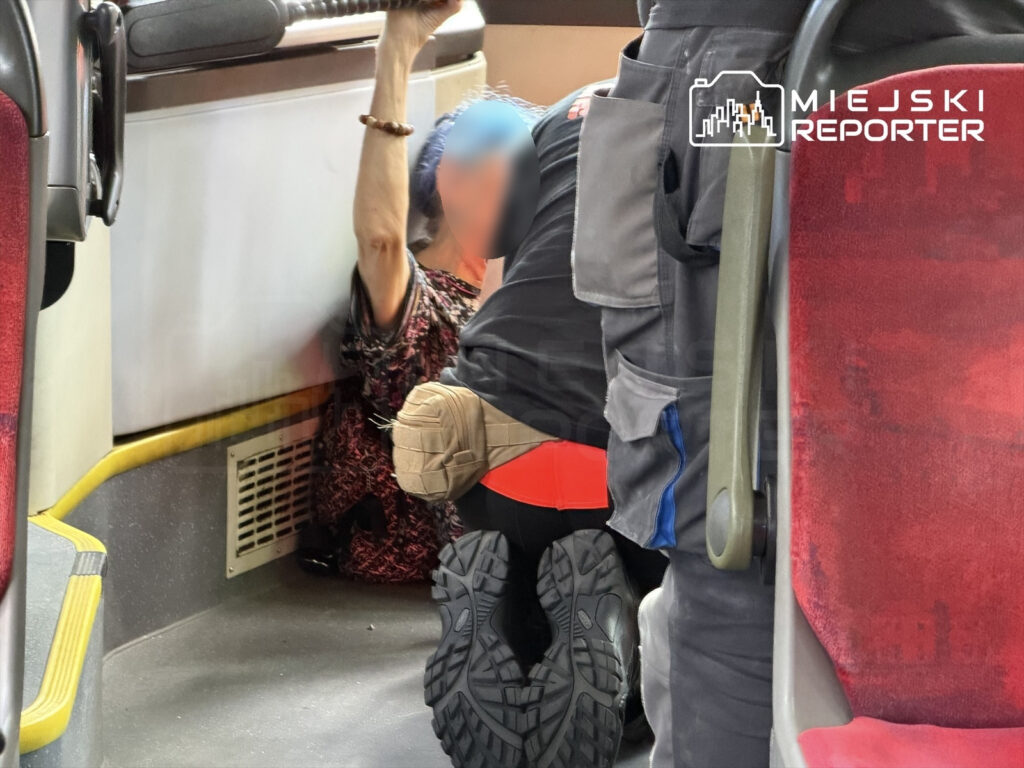 Kobieta z niebieskimi włosami trzyma się poręczy w autobusie, podczas gdy ratownik medyczny udziela jej pomocy.