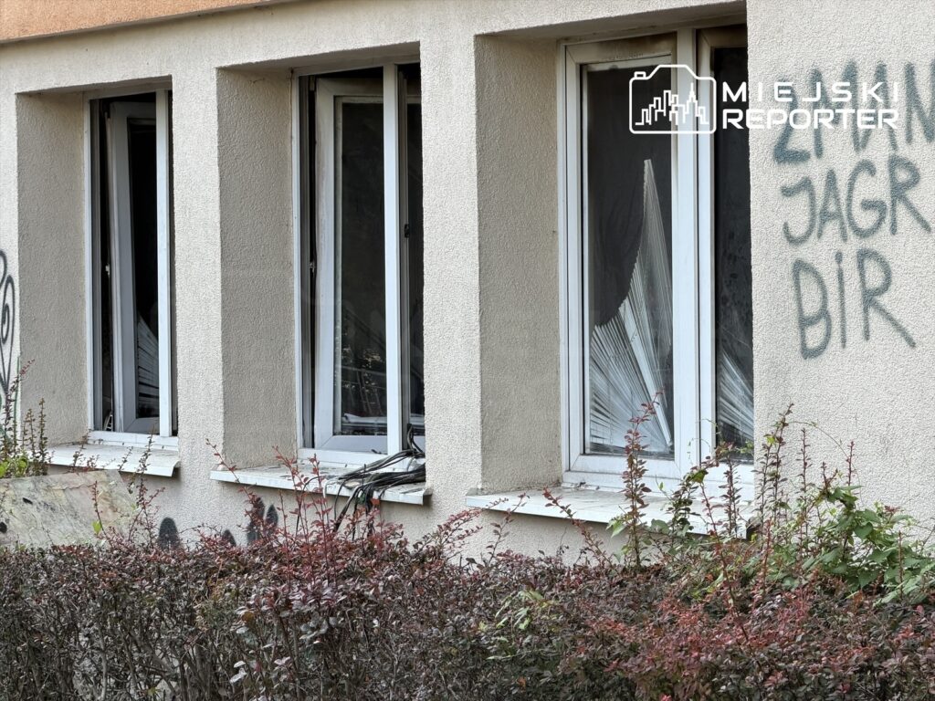 Uszkodzone okna w budynku z widocznymi śladami po pożarze, z graffiti na elewacji oraz zaroślami w tle.