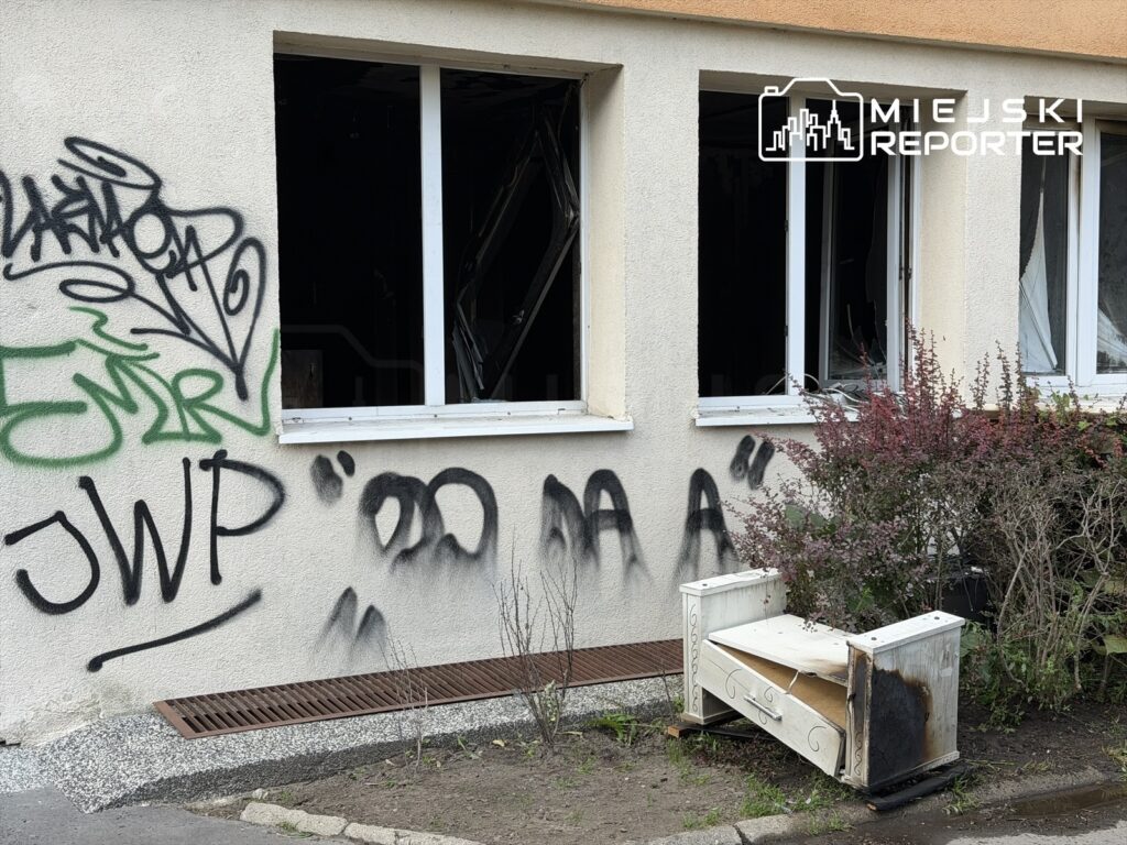 Zniszczona elewacja budynku z wybitymi oknami i graffiti, obok uszkodzony mebel leżący na chodniku.