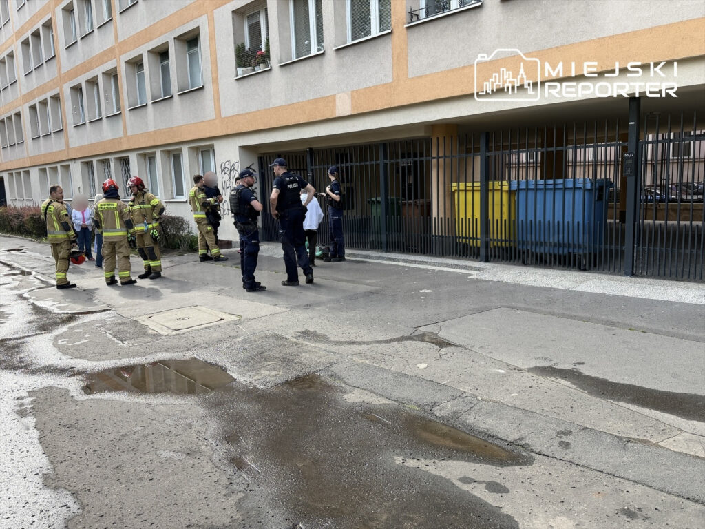 Grupa strażaków i policjantów rozmawia na chodniku przed budynkiem mieszkalnym, obok pojemników na odpady.
