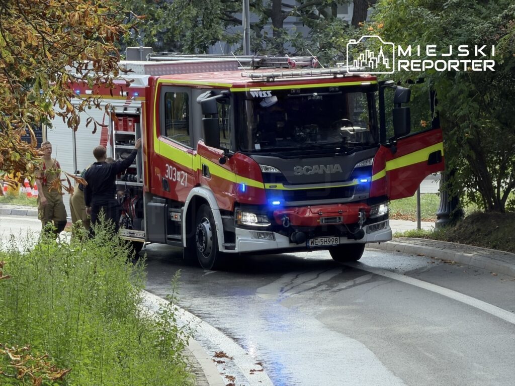 Czerwony wóz strażacki z włączonymi sygnałami świetlnymi stoi na zakręcie, a strażacy przygotowują sprzęt do akcji.