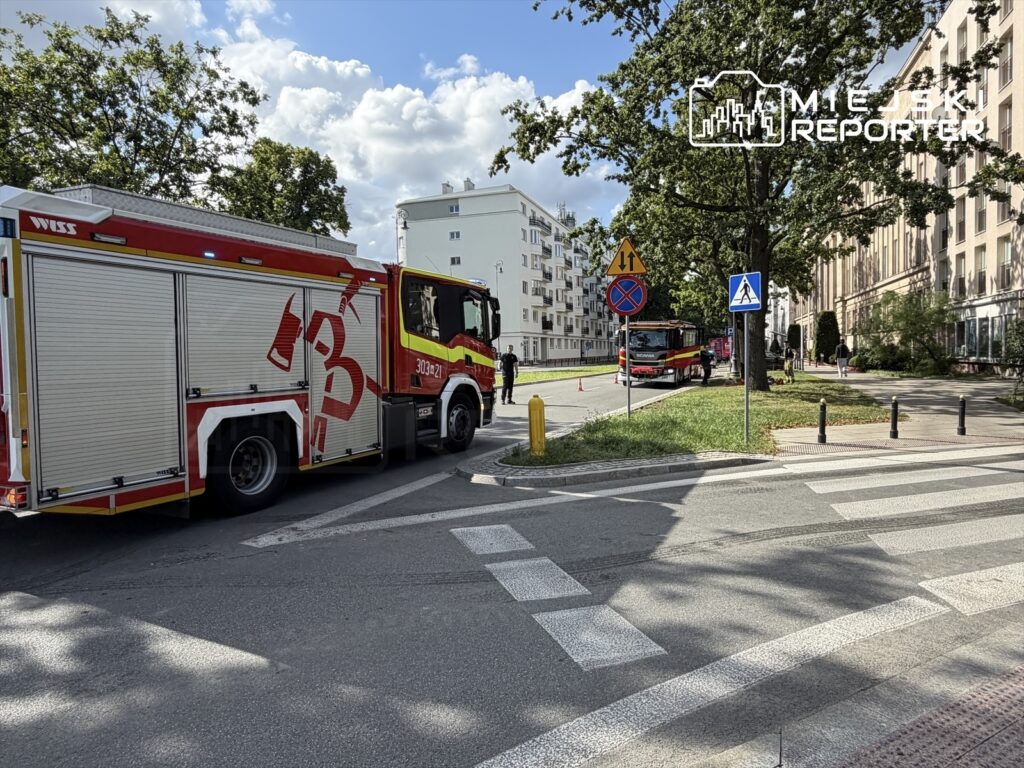 Wozidło strażackie zatrzymane na zakręcie ulicy, obok znaków drogowych i budynków mieszkalnych.