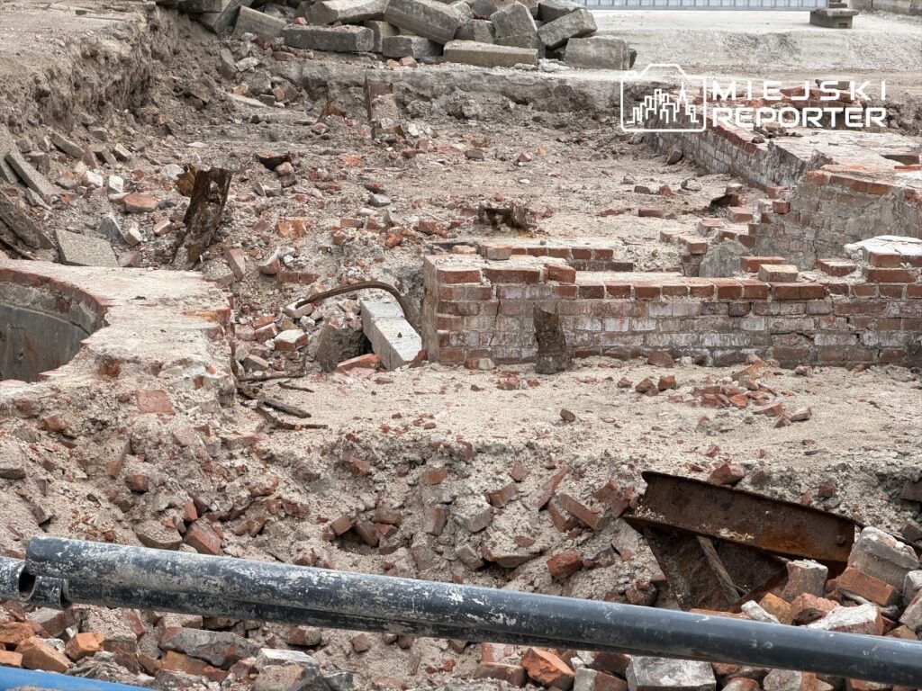 Fragmenty cegieł i zardzewiałe elementy metalowe w wykopie budowlanym, na tle gruzowiska i piasku.