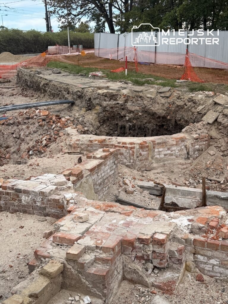 Wykop z ceglaną strukturą, odsłaniający fragment dawnej architektury, otoczony płotem budowlanym i ziemią.