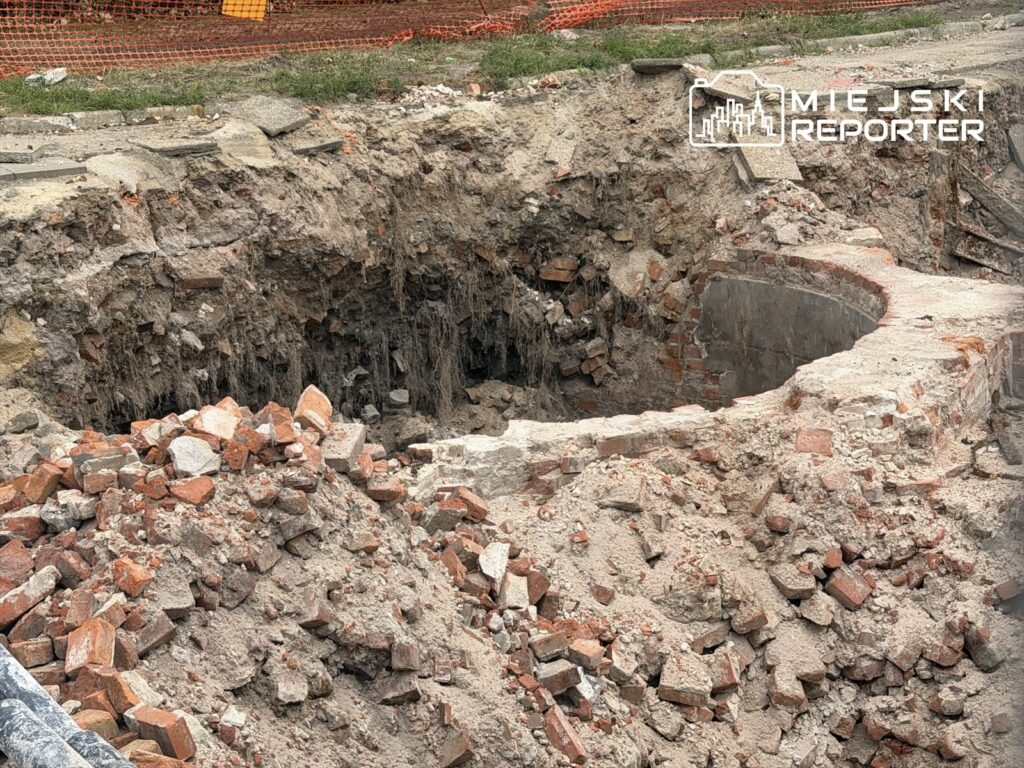 Wykop z fragmentami cegieł i gruzu, odsłaniający pozostałości dawnej struktury, otoczony pomarańczową siatką.