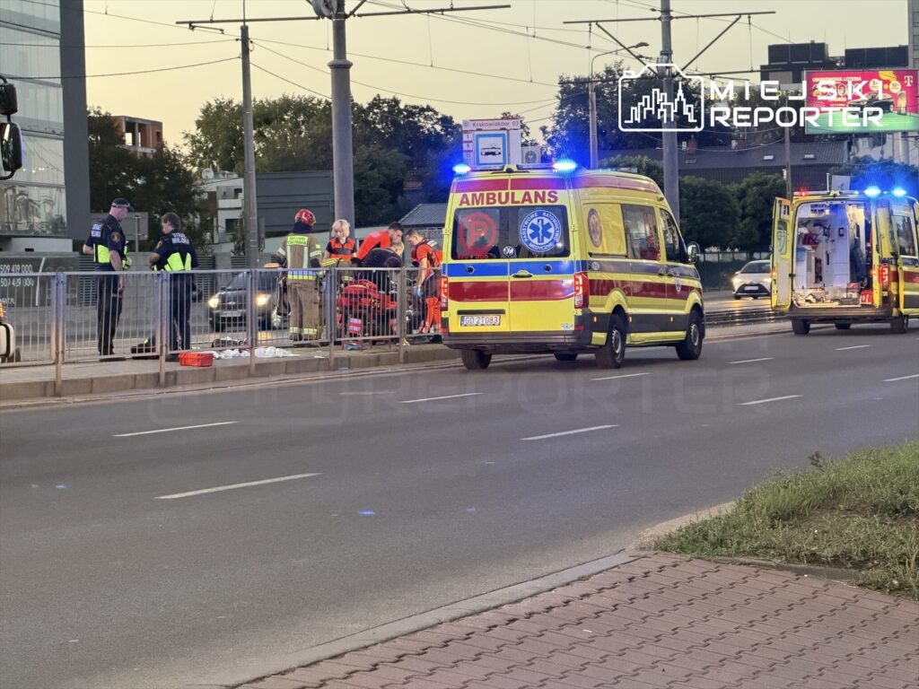 Grupa ratowników medycznych oraz policjantów udziela pomocy mężczyźnie leżącemu na chodniku przy przystanku.