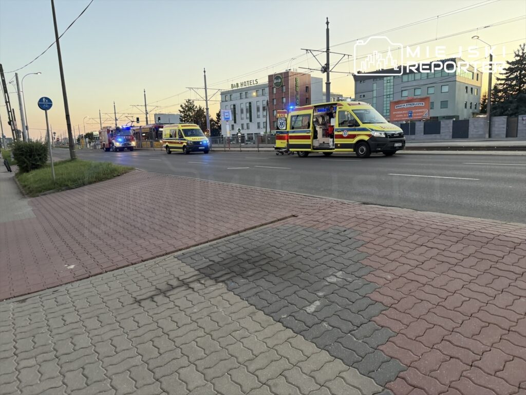Karetka pogotowia oraz wóz strażacki przejeżdżają obok przystanku tramwajowego, w tle widać nowoczesne budynki.
