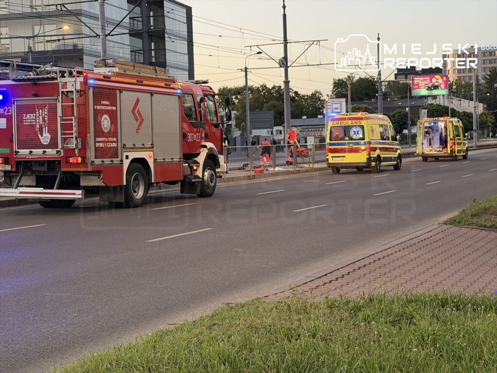 Wozidła strażackie i ambulansy zatrzymane przy przystanku, obok nowoczesnego biurowca, podczas akcji ratunkowej.