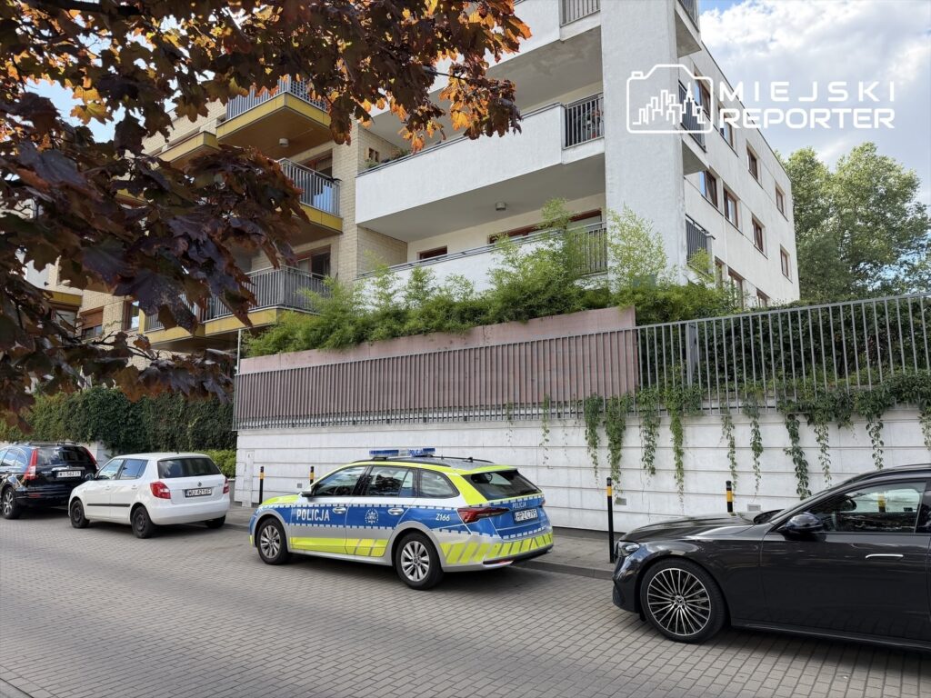 Policyjny radiowóz zaparkowany na ulicy przed nowoczesnym budynkiem mieszkalnym, otoczony przez samochody osobowe.