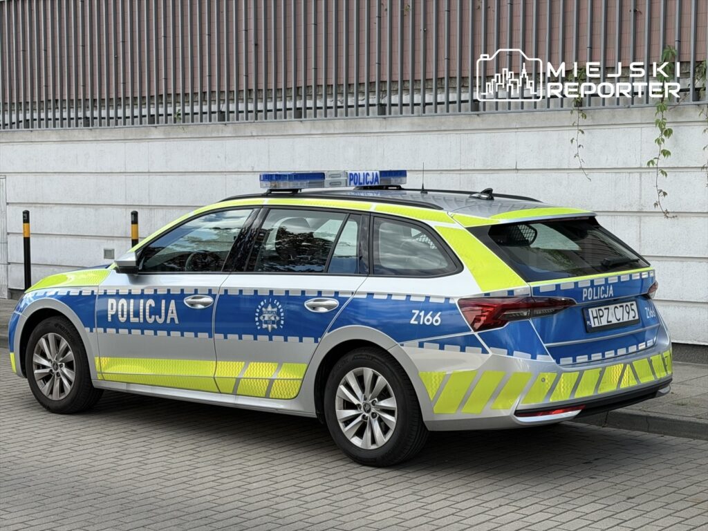 Radiowóz policyjny z niebiesko-żółtym oznakowaniem zaparkowany na chodniku obok budynku.