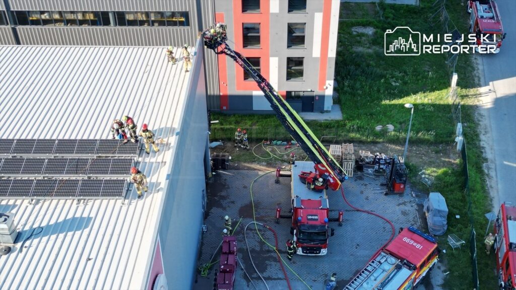 Strażacy na dachu hali, używający drabiny przysuwnej do gaszenia pożaru, w tle wozów strażackich i sprzętu.