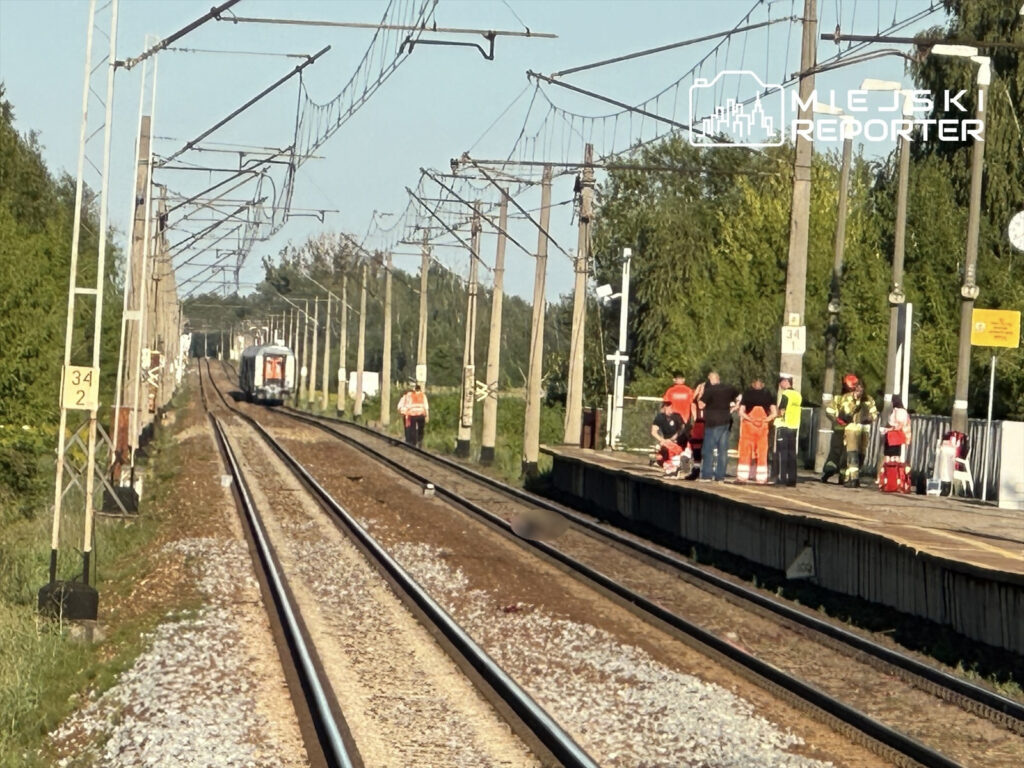 Na torach kolejowych widoczne są służby ratunkowe, w tym strażacy, oraz kilka osób na peronie. W tle nadjeżdża pociąg.