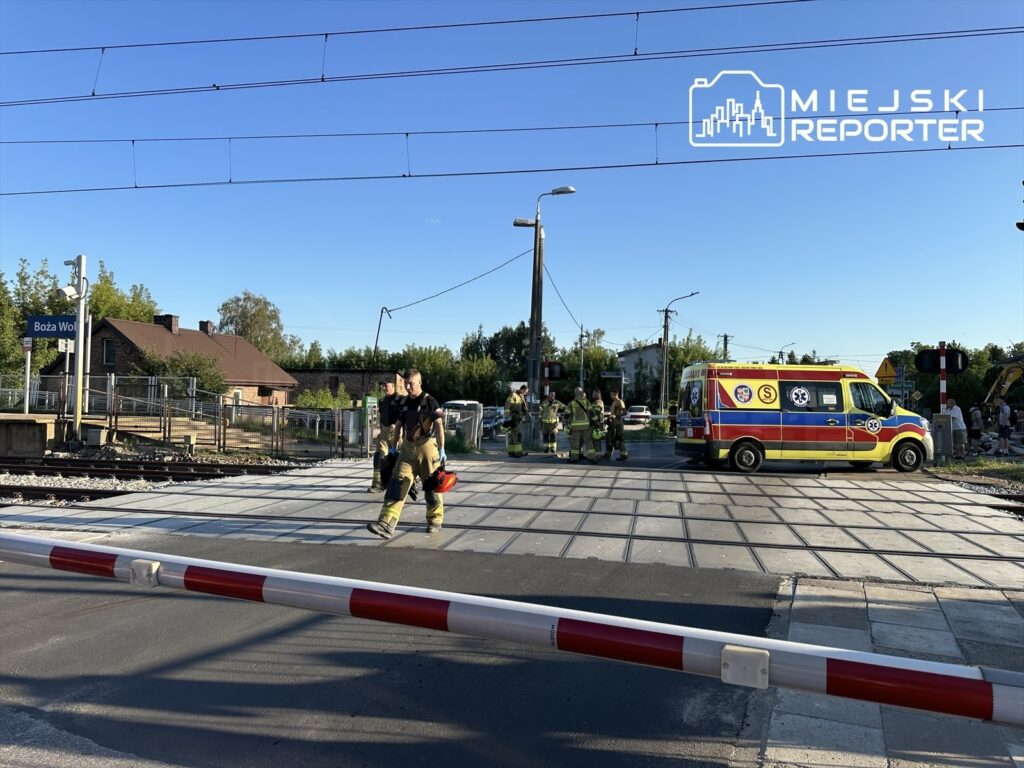 Ratownicy medyczni i strażacy wchodzą na przejazd kolejowy, obok stoi karetka pogotowia. W tle widoczny znak stacji.